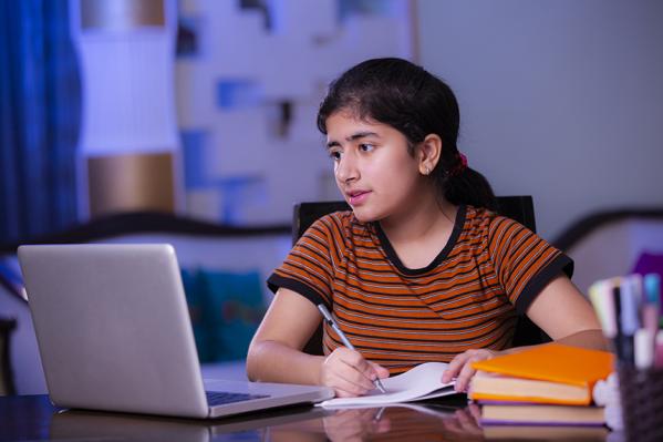 A teenager looking at a laptop and writing in a notepad. She has black hair.