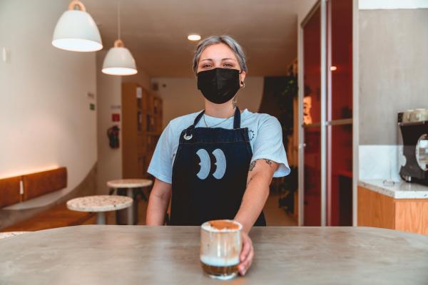 A young barista wearing a mask handing over a coffee