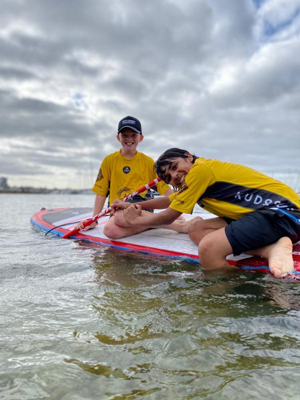 Two young Aboriginal people on a paddle board in the ocean, smiling